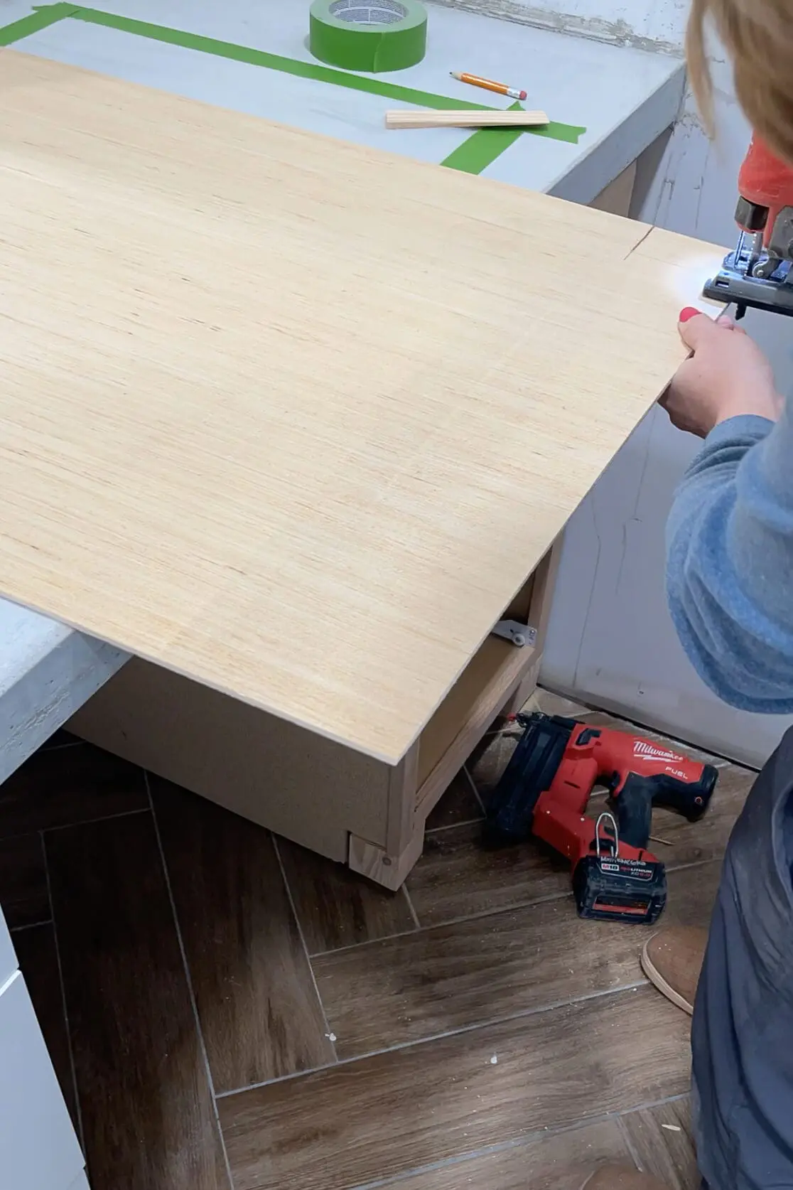 Person using a jigsaw to cut a wood veneer sheet placed over a countertop during a bathroom renovation. Tools, painter’s tape, and measuring supplies are visible on the workspace.